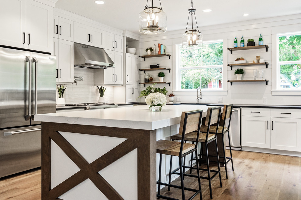 Inspiration for a country l-shaped kitchen in Portland with a farmhouse sink, shaker cabinets, white cabinets, white splashback, stainless steel appliances, medium hardwood floors, with island, brown floor and black benchtop.
