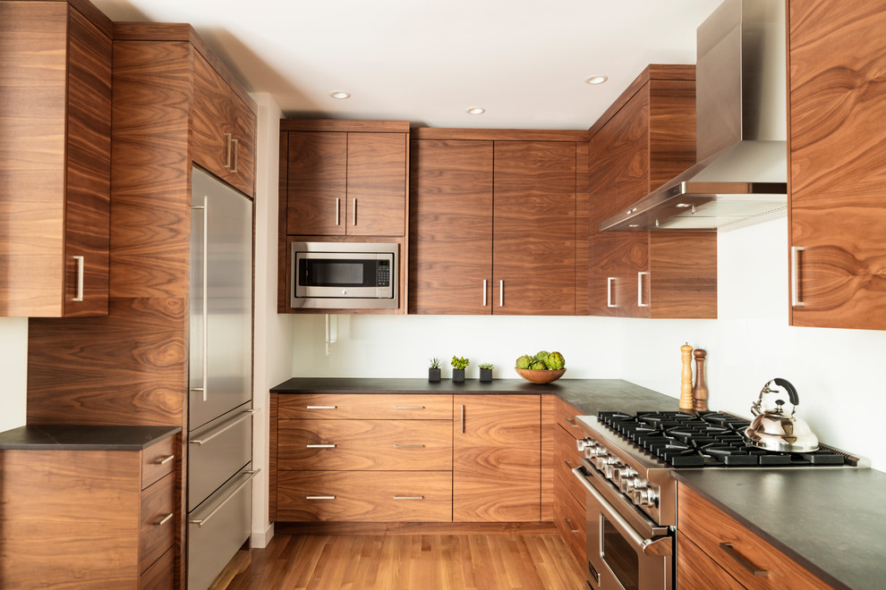 This is an example of a midcentury u-shaped eat-in kitchen in Seattle with an undermount sink, flat-panel cabinets, medium wood cabinets, quartz benchtops, white splashback, glass sheet splashback, stainless steel appliances, light hardwood floors, a peninsula, brown floor and black benchtop.