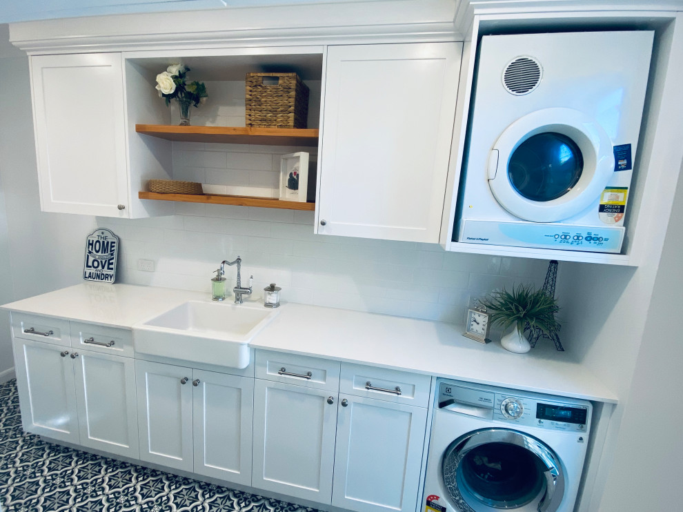 Inspiration for a large beach style galley dedicated laundry room in Brisbane with a farmhouse sink, shaker cabinets, white cabinets, quartz benchtops, white splashback, subway tile splashback, white walls, ceramic floors, black floor, white benchtop, recessed and panelled walls.