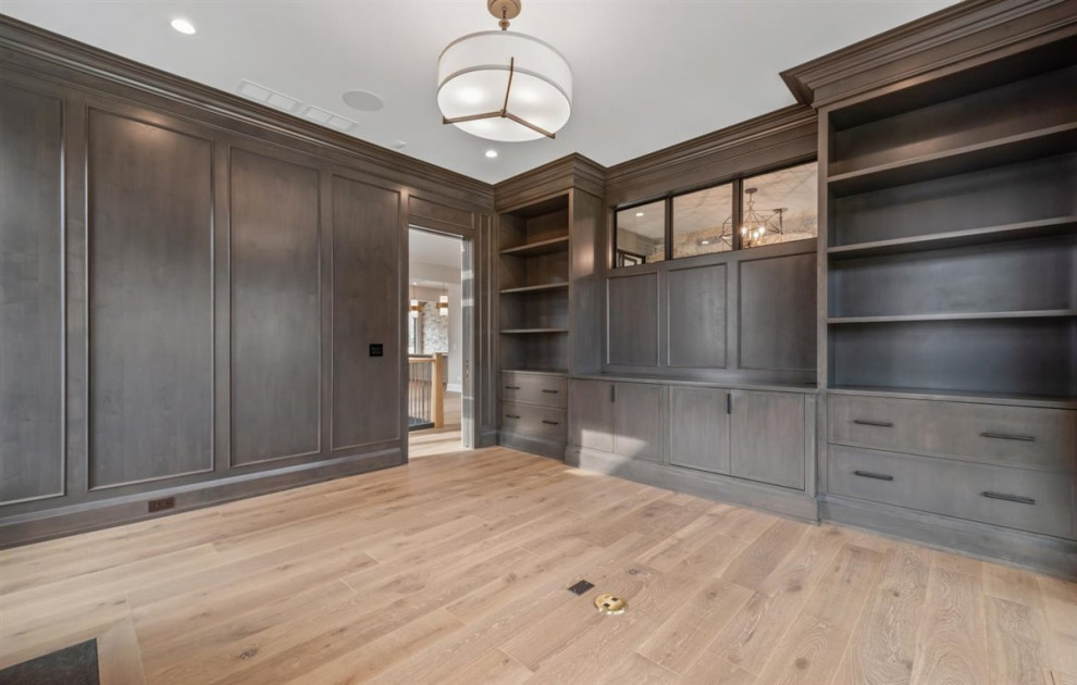 Large transitional home office in Chicago with a library, grey walls, light hardwood floors, a standard fireplace, a wood fireplace surround, a freestanding desk, brown floor and panelled walls.