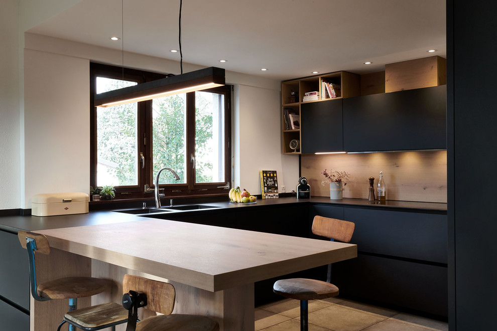 Design ideas for a large contemporary u-shaped open plan kitchen in Other with a double-bowl sink, flat-panel cabinets, black cabinets, timber splashback, black appliances, ceramic floors, black benchtop, a peninsula and beige floor.