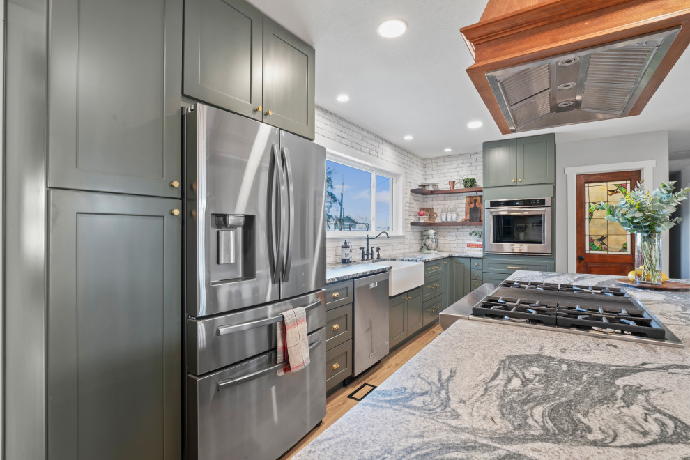 Bild på ett stort lantligt grå grått kök, med en rustik diskho, skåp i shakerstil, gröna skåp, granitbänkskiva, vitt stänkskydd, stänkskydd i tegel, rostfria vitvaror, vinylgolv, en köksö och beiget golv