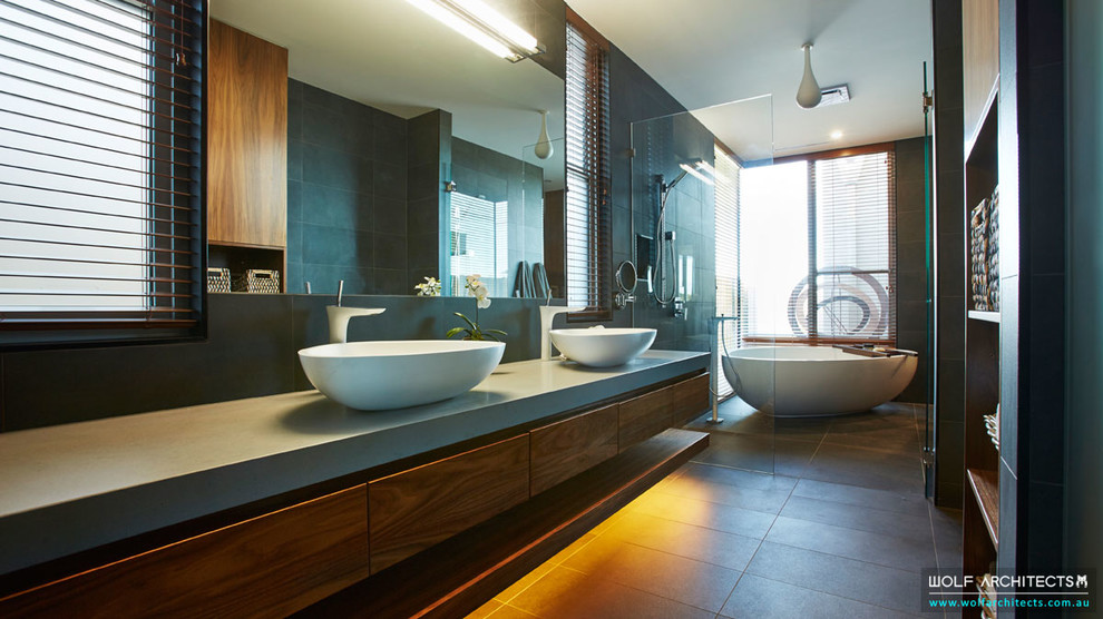Photo of a mid-sized contemporary master wet room bathroom in Melbourne with furniture-like cabinets, medium wood cabinets, a freestanding tub, a one-piece toilet, gray tile, ceramic tile, grey walls, porcelain floors, a console sink, engineered quartz benchtops, beige floor and a hinged shower door.