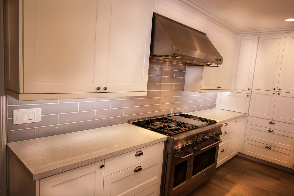 Example of a large transitional u-shaped light wood floor kitchen pantry design with a farmhouse sink, shaker cabinets, white cabinets, quartz countertops, gray backsplash, glass tile backsplash, stainless steel appliances and an island