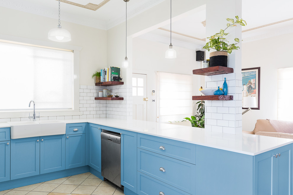 This is an example of a mid-sized transitional u-shaped kitchen pantry in Sydney with a farmhouse sink, shaker cabinets, blue cabinets, solid surface benchtops, white splashback, subway tile splashback, stainless steel appliances, terra-cotta floors, with island, beige floor and white benchtop.