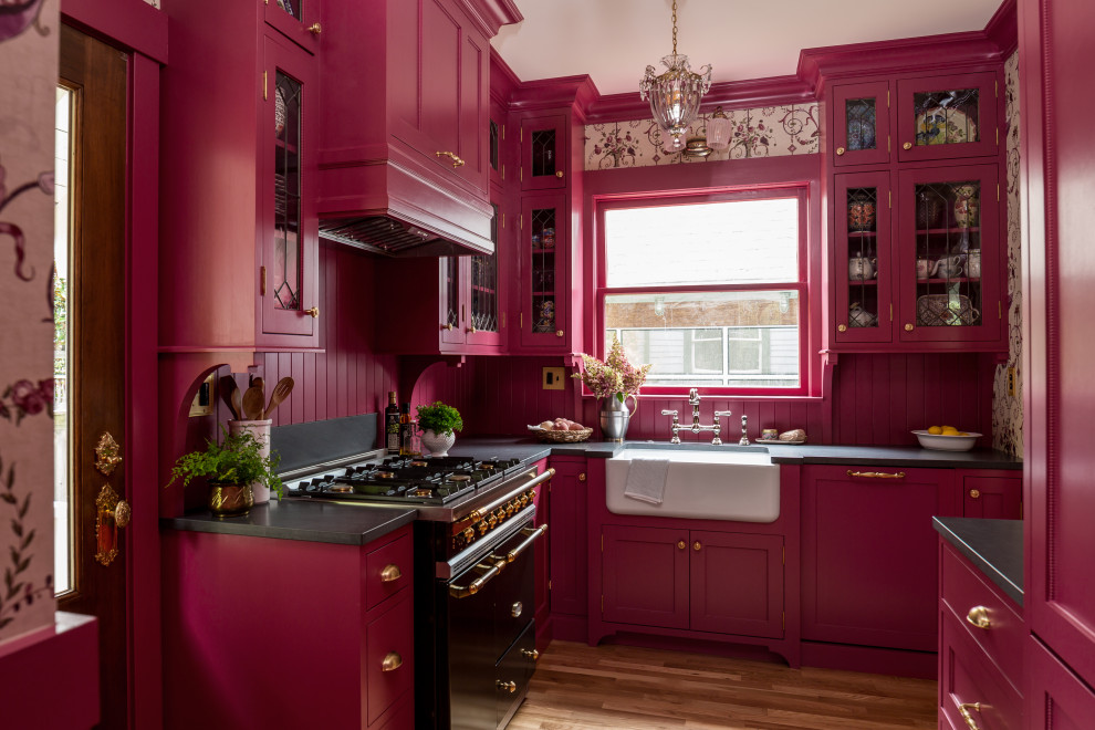 This is an example of a small traditional l-shaped enclosed kitchen in Seattle with a belfast sink, shaker cabinets, pink cabinets, granite worktops, pink splashback, wood splashback, integrated appliances, light hardwood flooring, no island, brown floors and black worktops.