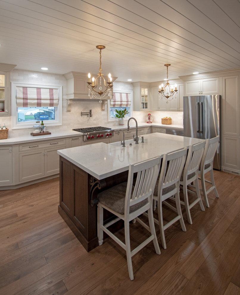 Example of a transitional medium tone wood floor, brown floor and shiplap ceiling kitchen design with a farmhouse sink, shaker cabinets, beige cabinets, quartz countertops, white backsplash, ceramic backsplash, stainless steel appliances, an island and white countertops
