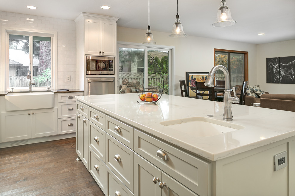 Design ideas for a mid-sized arts and crafts l-shaped eat-in kitchen in Portland with a farmhouse sink, flat-panel cabinets, white cabinets, quartzite benchtops, white splashback, subway tile splashback, stainless steel appliances, dark hardwood floors and with island.