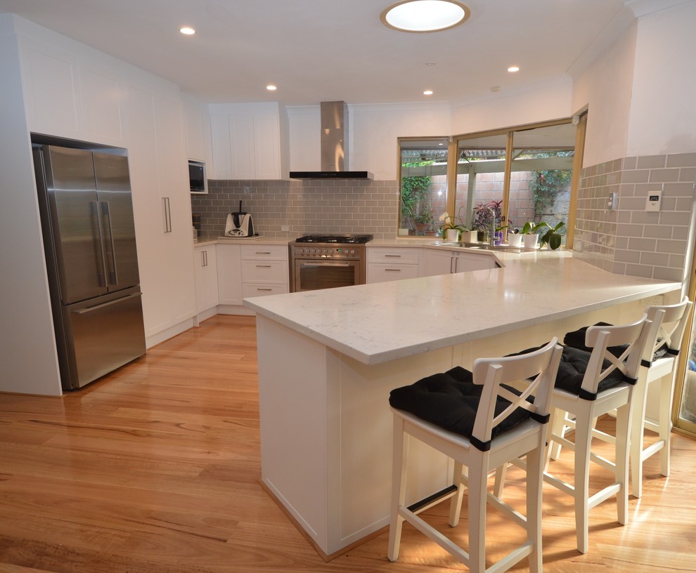 Large traditional u-shaped eat-in kitchen in Perth with a drop-in sink, recessed-panel cabinets, white cabinets, marble benchtops, grey splashback, ceramic splashback, stainless steel appliances, light hardwood floors, a peninsula and brown floor.