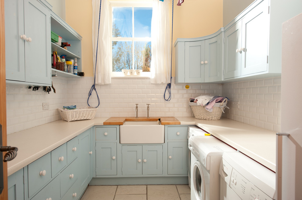 Mid-sized traditional u-shaped dedicated laundry room in London with a farmhouse sink, blue cabinets, solid surface benchtops, beige walls, a side-by-side washer and dryer and beige benchtop.