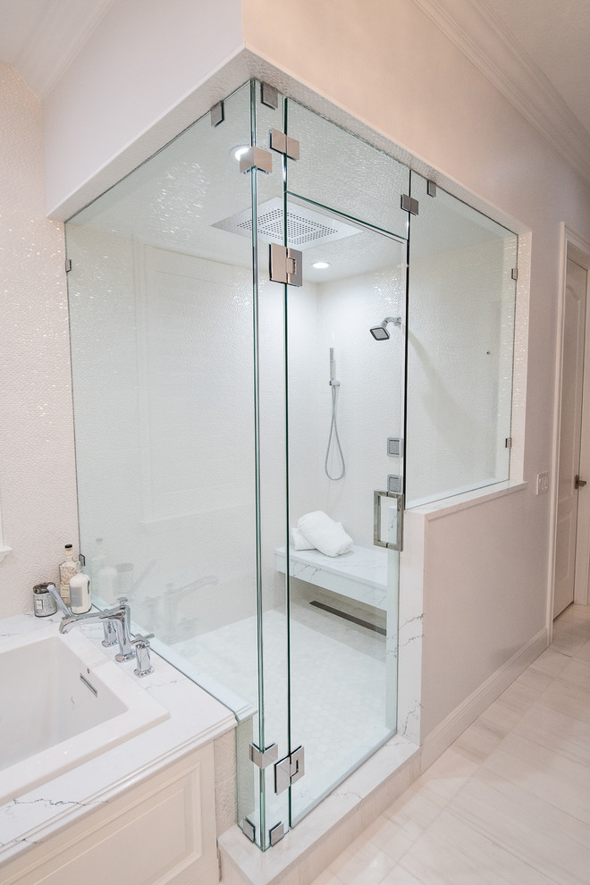 This is an example of a large transitional master bathroom in Tampa with raised-panel cabinets, white cabinets, a drop-in tub, a double shower, a one-piece toilet, white tile, porcelain tile, grey walls, marble floors, an undermount sink, glass benchtops, white floor, a hinged shower door and white benchtops.