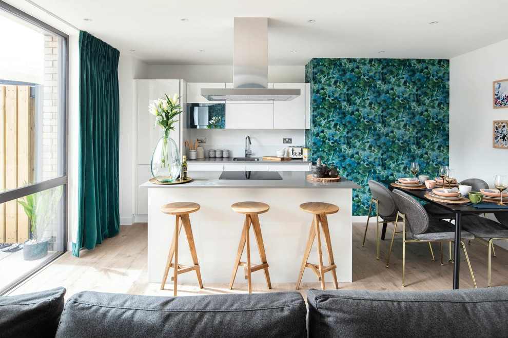 Mid-sized contemporary galley open plan kitchen in London with a drop-in sink, flat-panel cabinets, white cabinets, blue splashback, black appliances, light hardwood floors, with island, grey floor and grey benchtop.