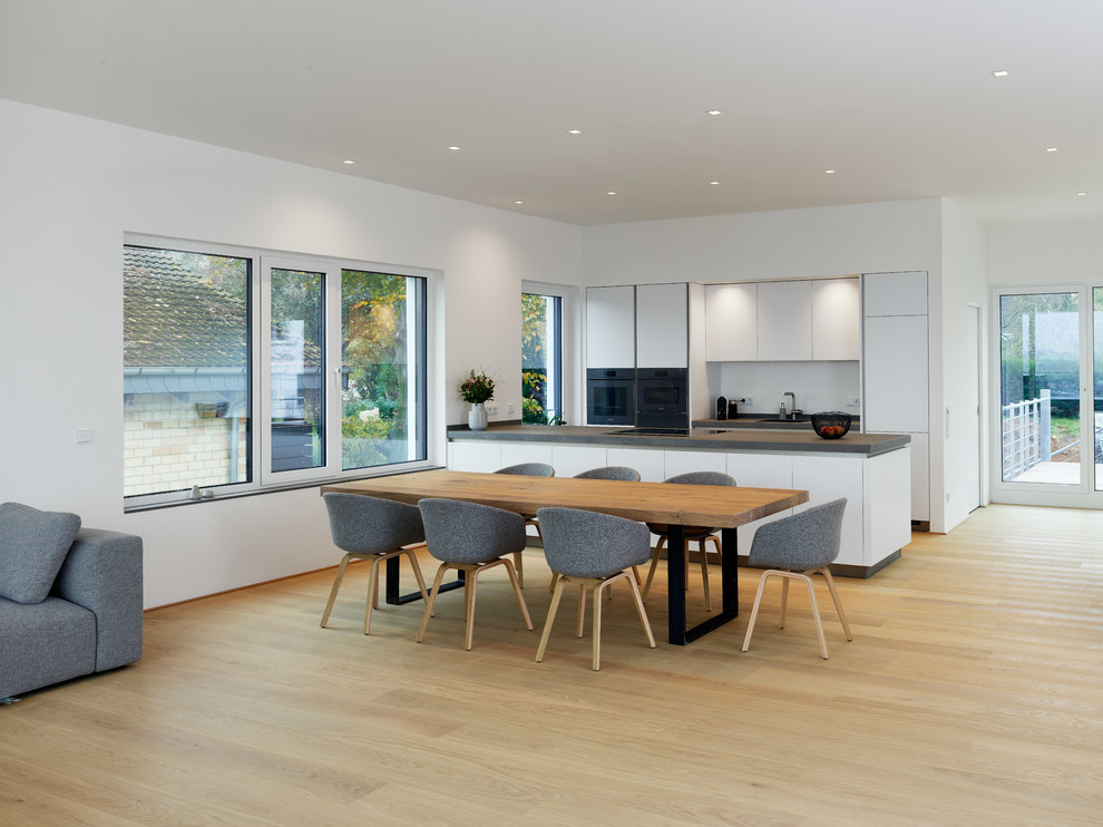 Design ideas for an expansive modern galley open plan kitchen in Other with flat-panel cabinets, white cabinets, white splashback, black appliances, light hardwood floors, with island, beige floor and grey benchtop.