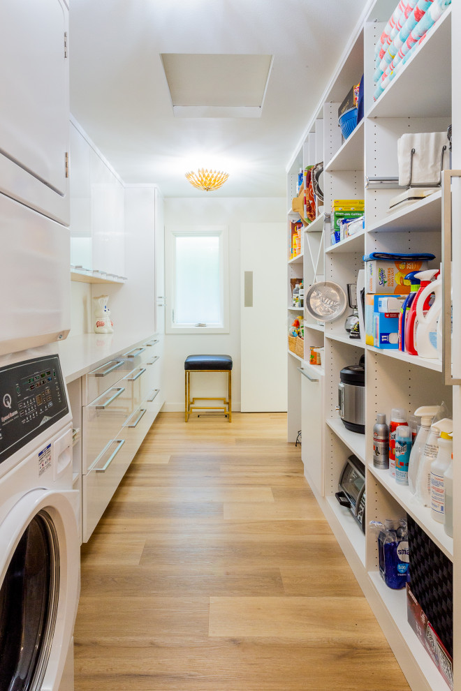 This is an example of a mid-sized contemporary galley dedicated laundry room in Seattle with flat-panel cabinets, white cabinets, quartzite benchtops, white walls, light hardwood floors, a stacked washer and dryer, brown floor and white benchtop.