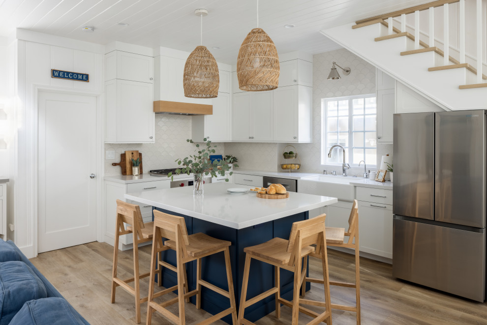 Photo of a transitional l-shaped kitchen in Orange County with a farmhouse sink, shaker cabinets, white cabinets, white splashback, stainless steel appliances, medium hardwood floors, with island, brown floor, white benchtop and timber.