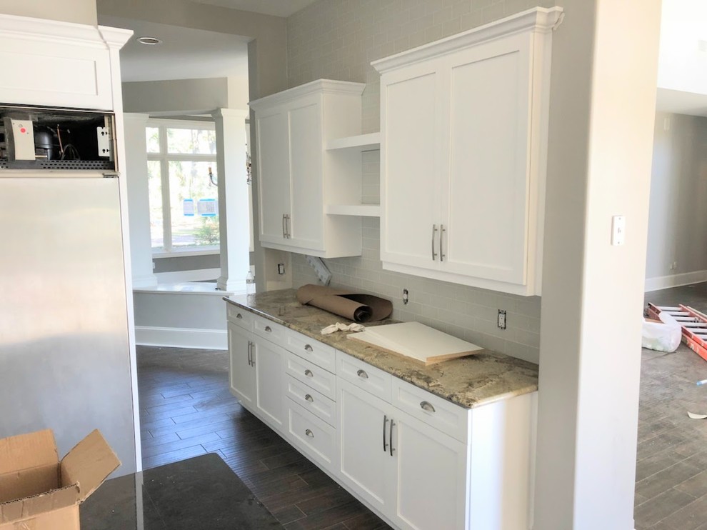 Inspiration for a mid-sized transitional porcelain tile and brown floor open concept kitchen remodel with a farmhouse sink, shaker cabinets, white cabinets, granite countertops, white backsplash, ceramic backsplash, stainless steel appliances and an island