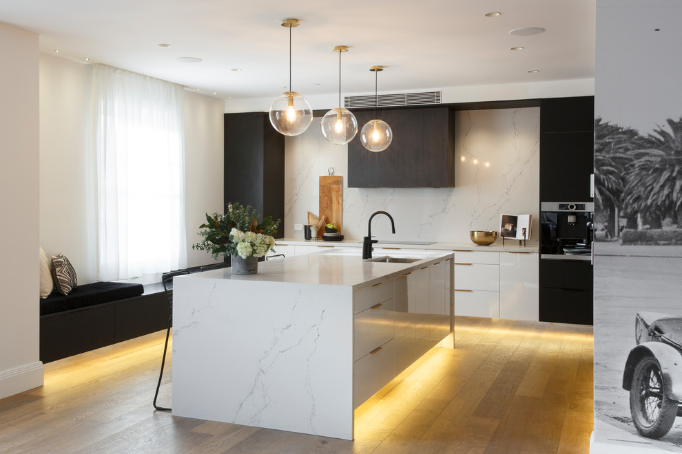 Photo of a contemporary l-shaped kitchen in Sydney with an undermount sink, flat-panel cabinets, white splashback, stone slab splashback, medium hardwood floors, with island, brown floor and white benchtop.