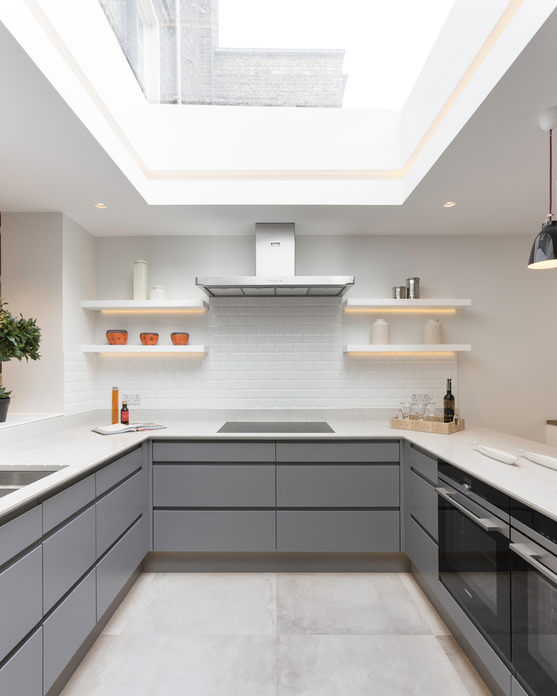 This is an example of a mid-sized contemporary u-shaped kitchen in London with a double-bowl sink, flat-panel cabinets, solid surface benchtops, white splashback, ceramic splashback, concrete floors, grey floor, grey cabinets, black appliances and a peninsula.