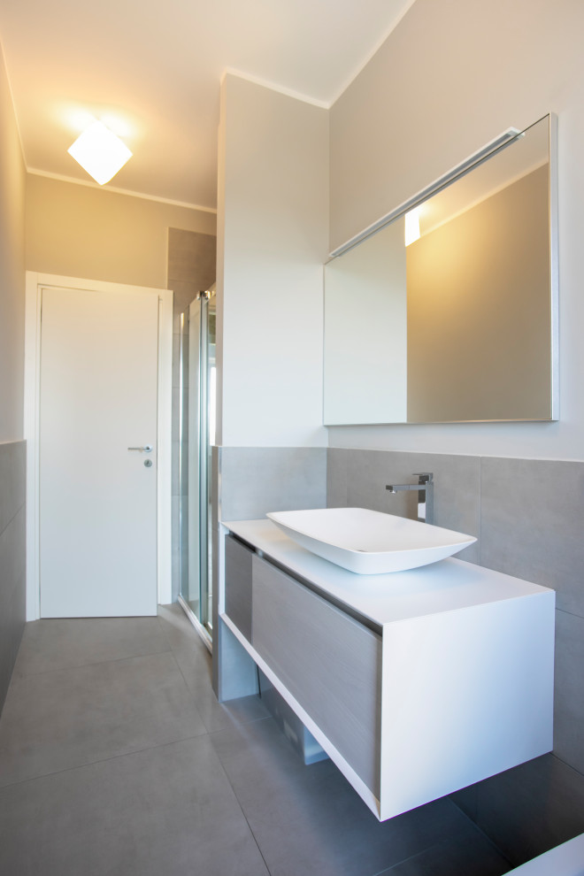 This is an example of a mid-sized contemporary 3/4 bathroom in Milan with beaded inset cabinets, light wood cabinets, an alcove shower, a wall-mount toilet, gray tile, porcelain tile, grey walls, painted wood floors, a vessel sink, solid surface benchtops, beige floor, a hinged shower door and white benchtops.