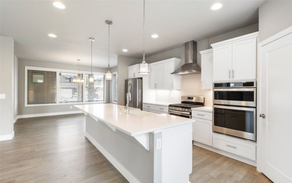 Photo of a large modern l-shaped eat-in kitchen in Portland with a farmhouse sink, recessed-panel cabinets, white cabinets, white splashback, stainless steel appliances, medium hardwood floors, with island and white benchtop.