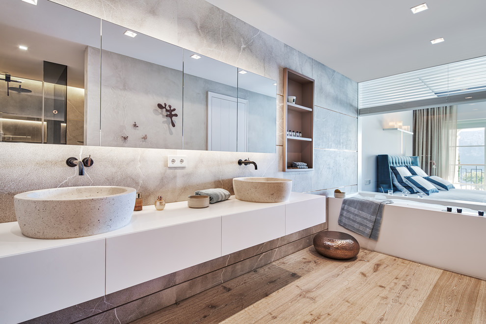 This is an example of a large contemporary master bathroom in Palma de Mallorca with flat-panel cabinets, white cabinets, gray tile, grey walls, medium hardwood floors, a vessel sink, brown floor, white benchtops and a drop-in tub.