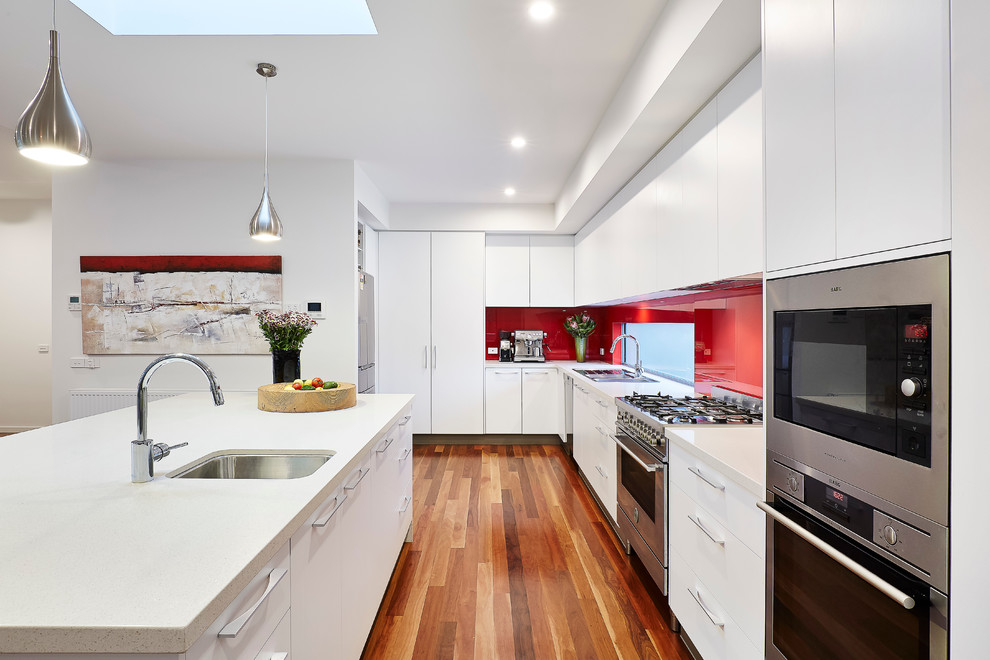 This is an example of a mid-sized modern l-shaped kitchen pantry in Melbourne with a double-bowl sink, recessed-panel cabinets, white cabinets, granite benchtops, red splashback, glass sheet splashback, stainless steel appliances, medium hardwood floors and with island.