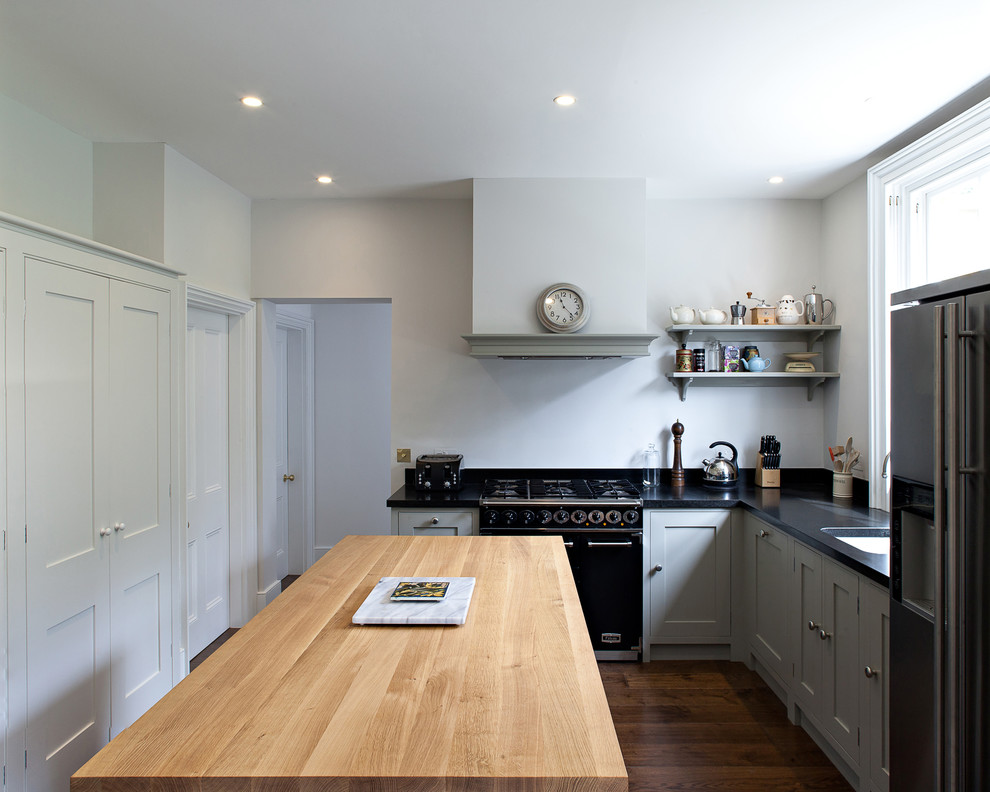 Mid-sized traditional l-shaped separate kitchen in London with a farmhouse sink, shaker cabinets, green cabinets, granite benchtops, black appliances, dark hardwood floors, with island and black splashback.