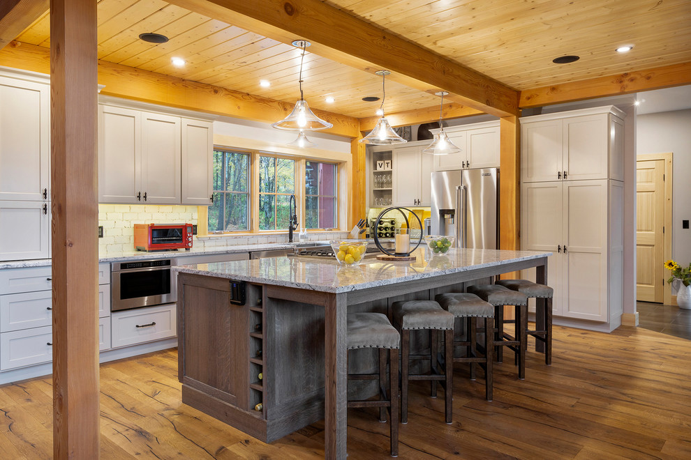 Inspiration for a large country l-shaped open plan kitchen in Boston with a farmhouse sink, shaker cabinets, granite benchtops, white splashback, stainless steel appliances, medium hardwood floors, with island, grey cabinets, brown floor and multi-coloured benchtop.