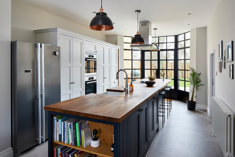 This is an example of a transitional galley separate kitchen in London with a farmhouse sink, shaker cabinets, white cabinets, wood benchtops, stainless steel appliances, concrete floors, with island, grey floor and brown benchtop.