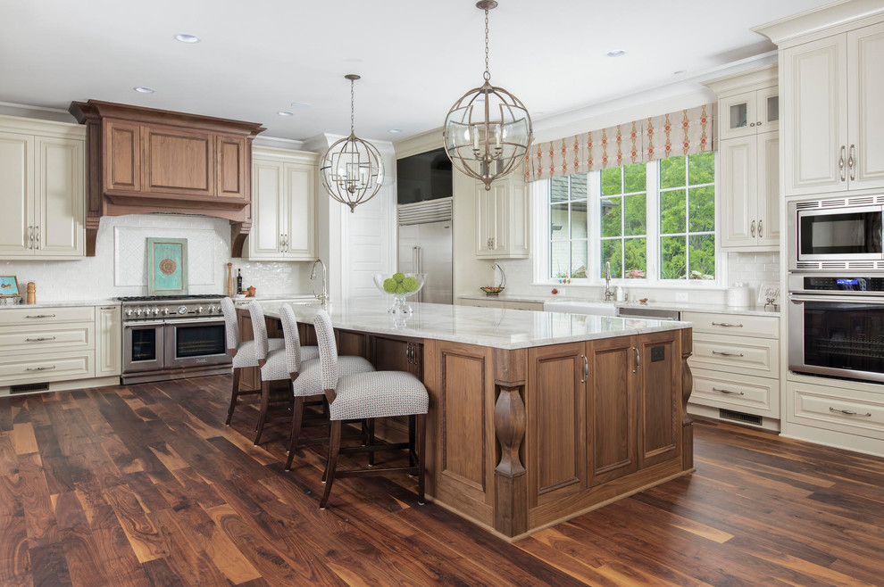 Inspiration for a large country l-shaped eat-in kitchen in Charlotte with a farmhouse sink, raised-panel cabinets, white cabinets, marble benchtops, white splashback, subway tile splashback, stainless steel appliances, medium hardwood floors, with island and brown floor.