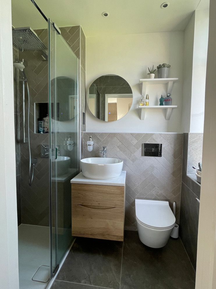 This is an example of a small modern master bathroom in London with flat-panel cabinets, medium wood cabinets, a curbless shower, a wall-mount toilet, gray tile, porcelain tile, a wall-mount sink, marble benchtops, a sliding shower screen, white benchtops, a single vanity and a floating vanity.