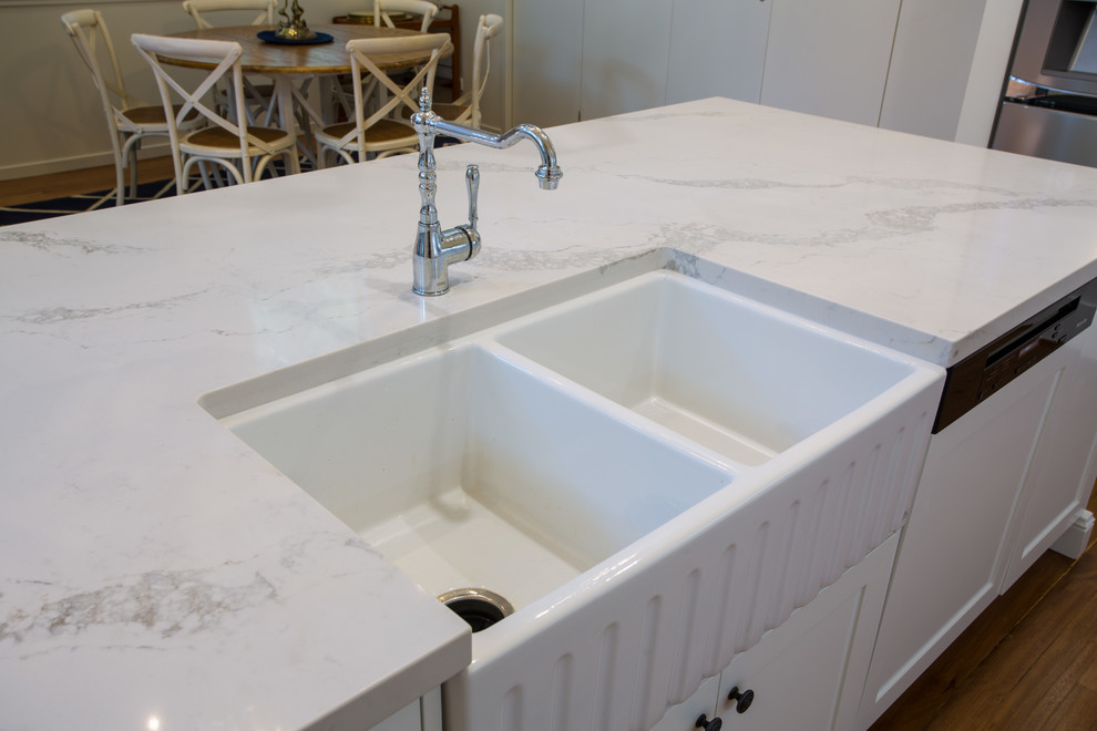 Inspiration for a large traditional galley kitchen pantry in Brisbane with a farmhouse sink, shaker cabinets, white cabinets, quartz benchtops, white splashback, subway tile splashback, white appliances, medium hardwood floors, with island, brown floor and white benchtop.