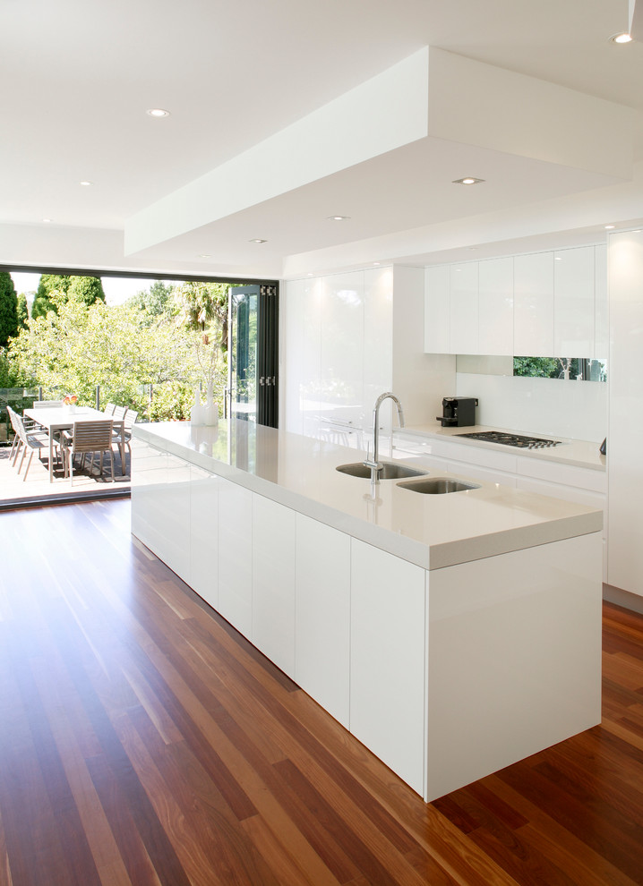 Photo of a modern galley eat-in kitchen in Sydney with a drop-in sink, flat-panel cabinets, white cabinets, quartz benchtops, white splashback, glass tile splashback, stainless steel appliances, medium hardwood floors and with island.