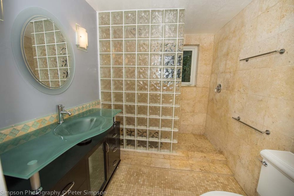 Photo of a mid-sized beach style master bathroom in Miami with a console sink, furniture-like cabinets, dark wood cabinets, glass benchtops, an open shower, a two-piece toilet, beige tile, multi-coloured walls and mosaic tile floors.