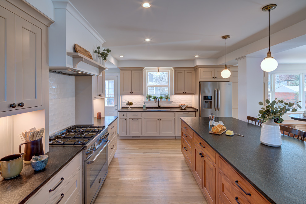 This is an example of a mid-sized country l-shaped open plan kitchen in DC Metro with a farmhouse sink, recessed-panel cabinets, beige cabinets, granite benchtops, white splashback, subway tile splashback, stainless steel appliances, light hardwood floors, with island and black benchtop.