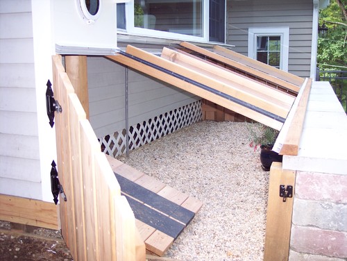 Enclosed dog run with a cover on the side of a house with a ramp coming out of a dog door. 
