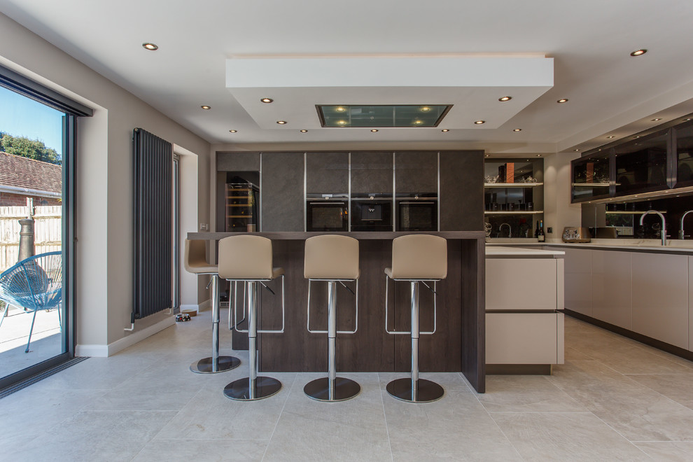 Contemporary l-shaped open plan kitchen in Sussex with flat-panel cabinets, mirror splashback, black appliances, with island, dark wood cabinets, beige floor and white benchtop.
