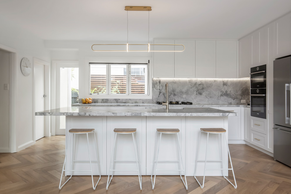 Large beach style l-shaped open plan kitchen in Sydney with an undermount sink, shaker cabinets, white cabinets, marble benchtops, grey splashback, marble splashback, stainless steel appliances, light hardwood floors, with island, multi-coloured floor, grey benchtop and coffered.