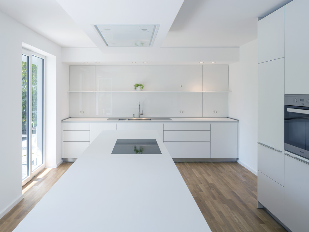 Design ideas for a large contemporary separate kitchen in Cologne with flat-panel cabinets, white cabinets, white splashback, glass sheet splashback, medium hardwood floors, with island, a drop-in sink and panelled appliances.