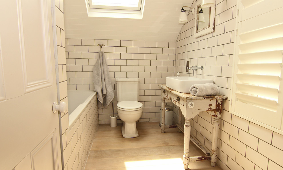 This is an example of a small beach style bathroom in Cornwall with furniture-like cabinets, distressed cabinets, an alcove tub, a shower/bathtub combo, a one-piece toilet, white tile, ceramic tile, white walls, light hardwood floors, a trough sink and wood benchtops.