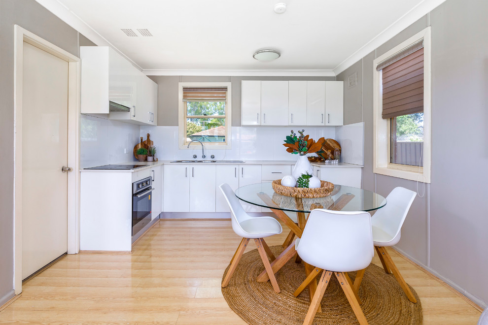 This is an example of a transitional l-shaped eat-in kitchen in Sydney with a double-bowl sink, flat-panel cabinets, white cabinets, white splashback, black appliances, light hardwood floors, beige floor and beige benchtop.