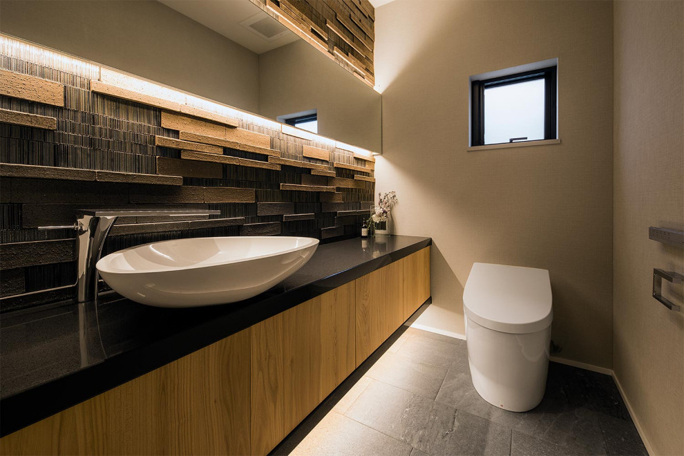Photo of a powder room with light wood cabinets, a one-piece toilet, brown tile, beige walls, a vessel sink, grey floor and black benchtops.