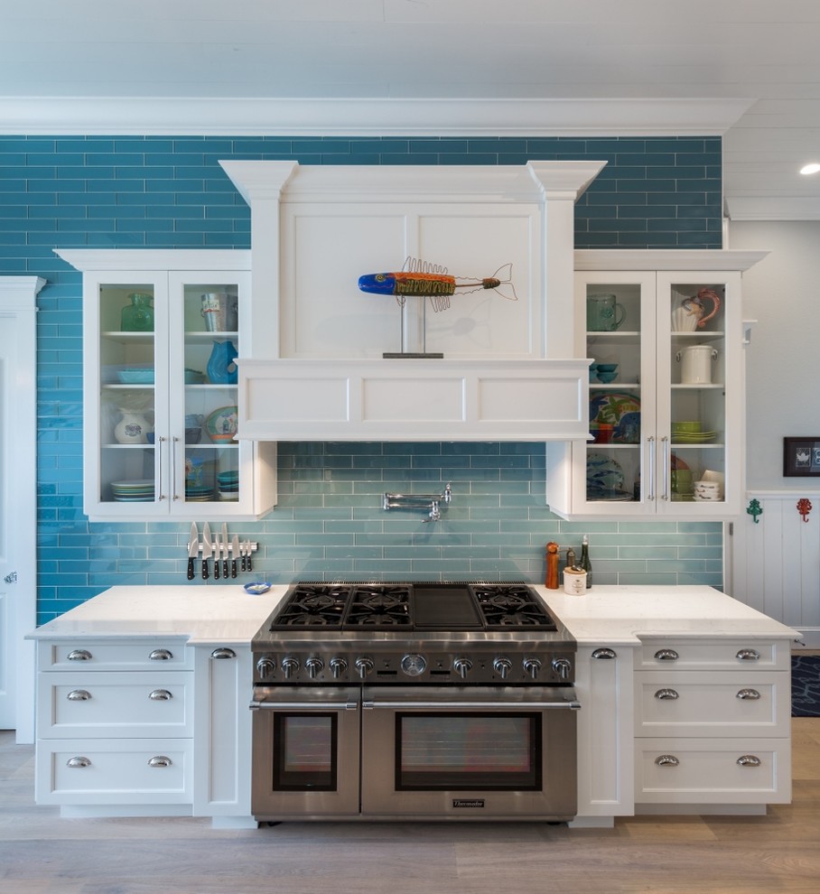 Photo of a mid-sized beach style galley eat-in kitchen in Tampa with a farmhouse sink, raised-panel cabinets, white cabinets, solid surface benchtops, blue splashback, subway tile splashback, stainless steel appliances, light hardwood floors and with island.