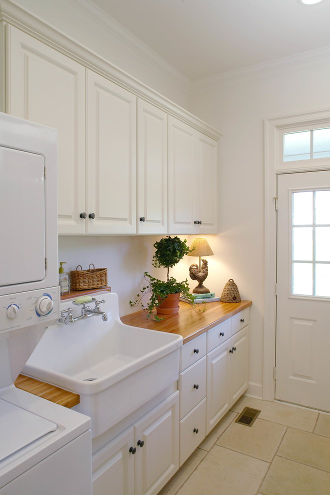 Design ideas for a small traditional laundry room in DC Metro with raised-panel cabinets, white cabinets, wood benchtops, white walls, a stacked washer and dryer, travertine floors, beige benchtop and a farmhouse sink.