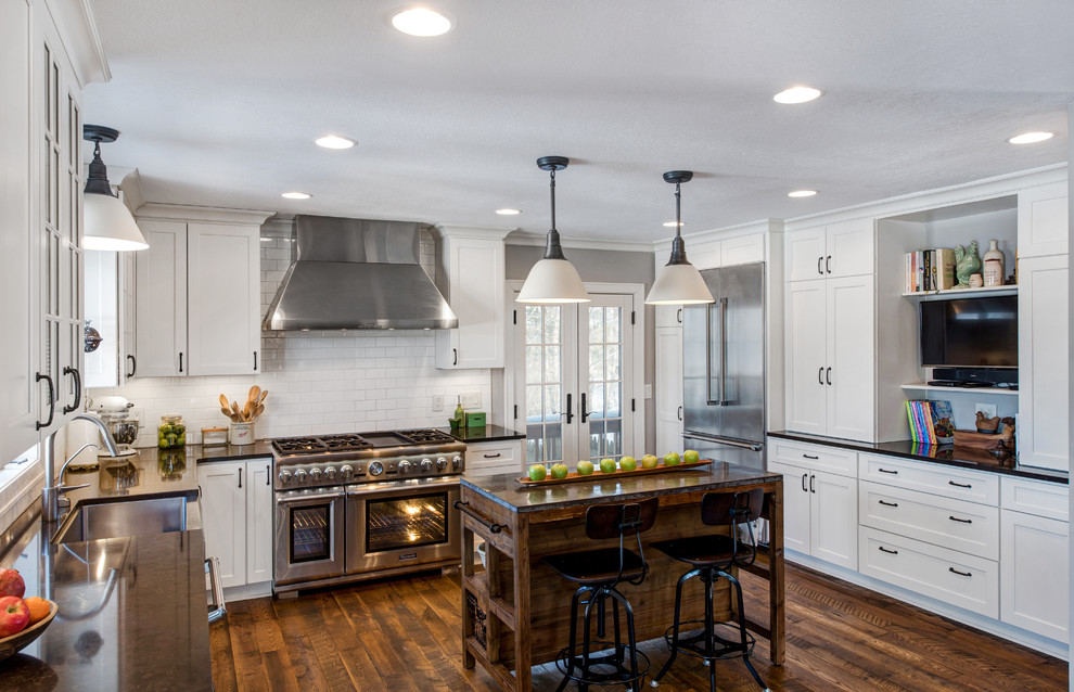 This is an example of a country l-shaped kitchen in Minneapolis with a farmhouse sink, shaker cabinets, white cabinets, white splashback, subway tile splashback, stainless steel appliances, dark hardwood floors, with island and black benchtop.