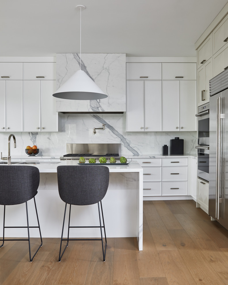 Large contemporary l-shaped eat-in kitchen in Toronto with an undermount sink, white cabinets, marble benchtops, white splashback, marble splashback, stainless steel appliances, medium hardwood floors, with island, brown floor and white benchtop.