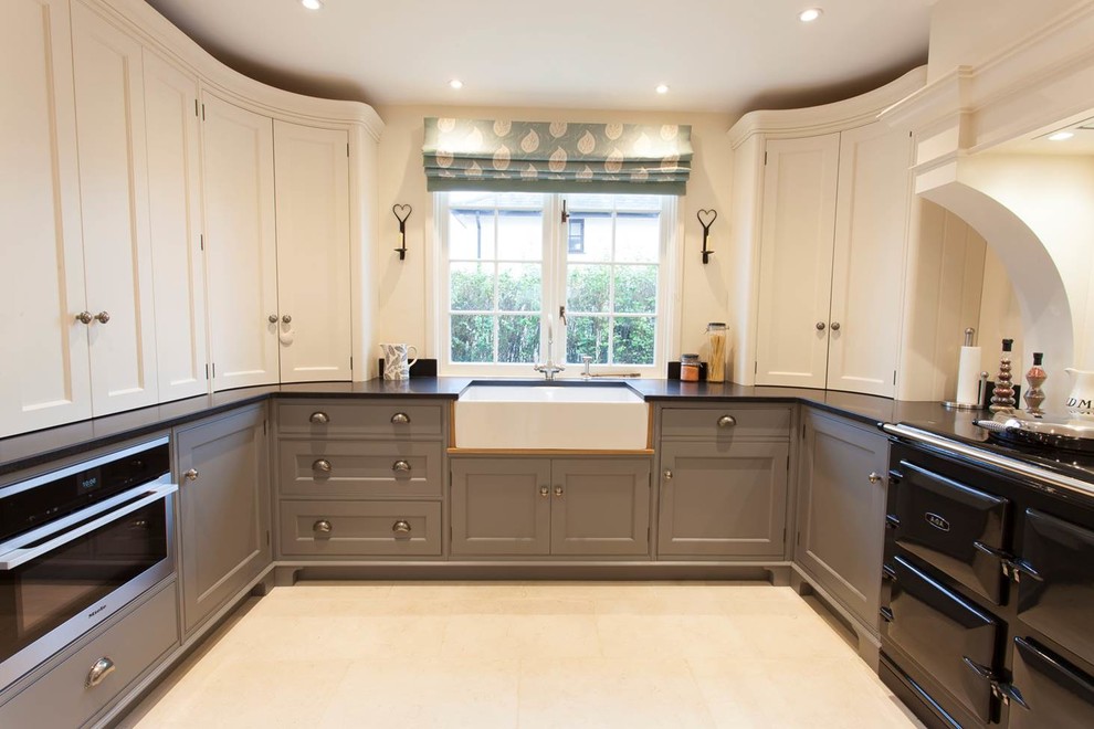 Mid-sized traditional u-shaped eat-in kitchen in Essex with a farmhouse sink, grey cabinets, granite benchtops, black appliances and limestone floors.