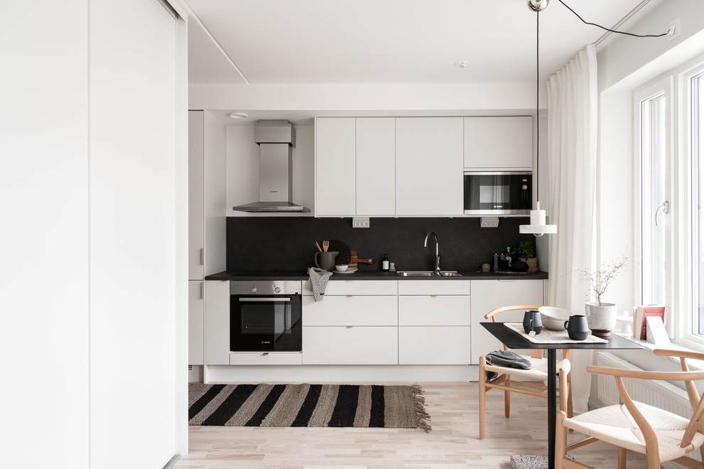 Design ideas for a small scandinavian single-wall eat-in kitchen in Gothenburg with a double-bowl sink, flat-panel cabinets, white cabinets, black splashback, slate splashback, panelled appliances, light hardwood floors, no island, beige floor and black benchtop.