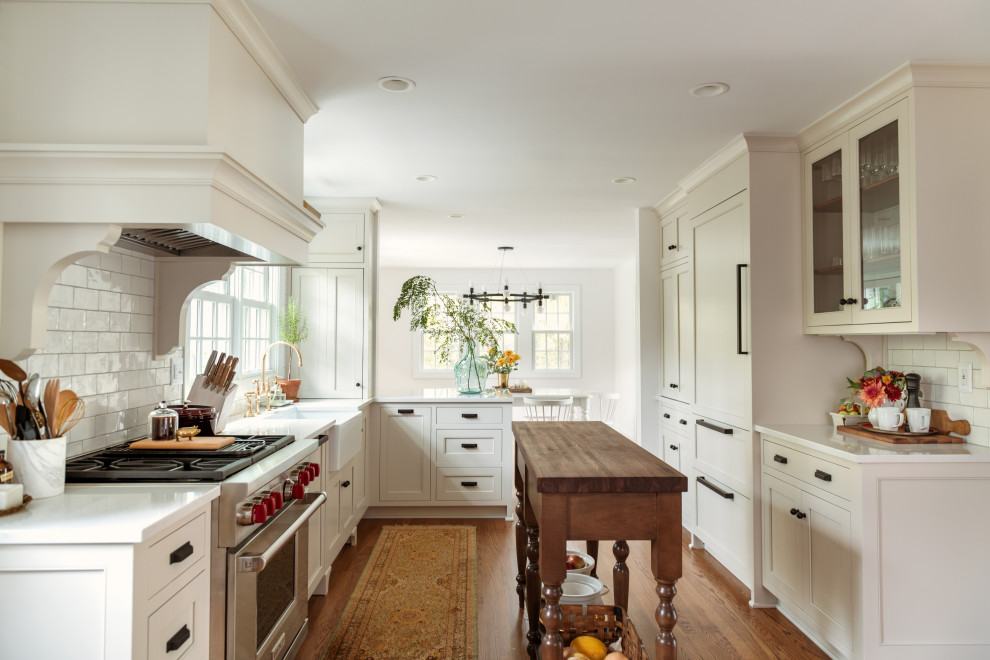 Photo of a traditional u-shaped kitchen in Other with a farmhouse sink, shaker cabinets, beige cabinets, white splashback, subway tile splashback, panelled appliances, medium hardwood floors, with island, brown floor and white benchtop.