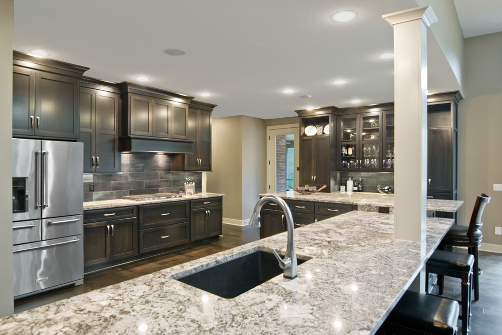 Large transitional u-shaped dark wood floor and brown floor open concept kitchen photo with an undermount sink, shaker cabinets, brown cabinets, granite countertops, black backsplash, porcelain backsplash, stainless steel appliances, an island and brown countertops